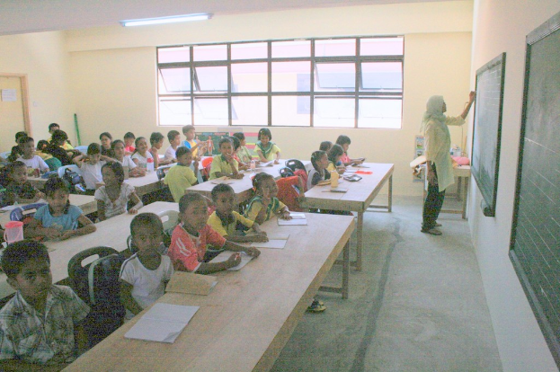 Ein Klassenzimmer mit einer Frau, die rechts an einer Tafel schreibt, Fenstern in der Mitte und einer Lampe oben, mit Tischen voller Bücher und Wasserflaschen und Stühlen mit Kindern in der Nähe.