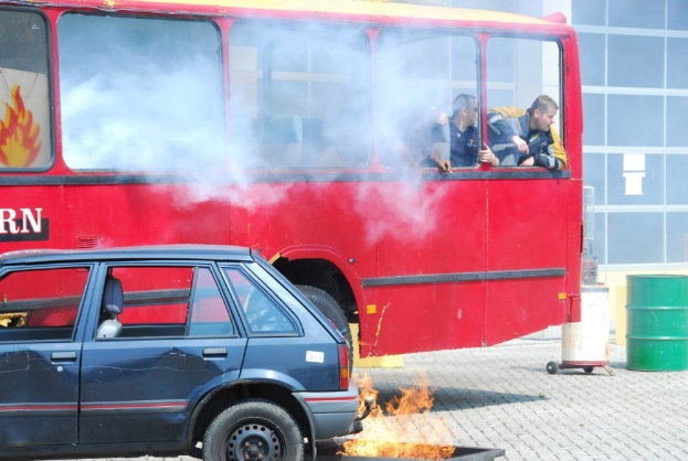 Roter Doppeldeckerbus mit Rauchentwicklung und drei sichtbaren Passagieren, neben einem Auto geparkt, vor einem Gebäude mit Glasfenstern und einem Fass auf der rechten Seite.