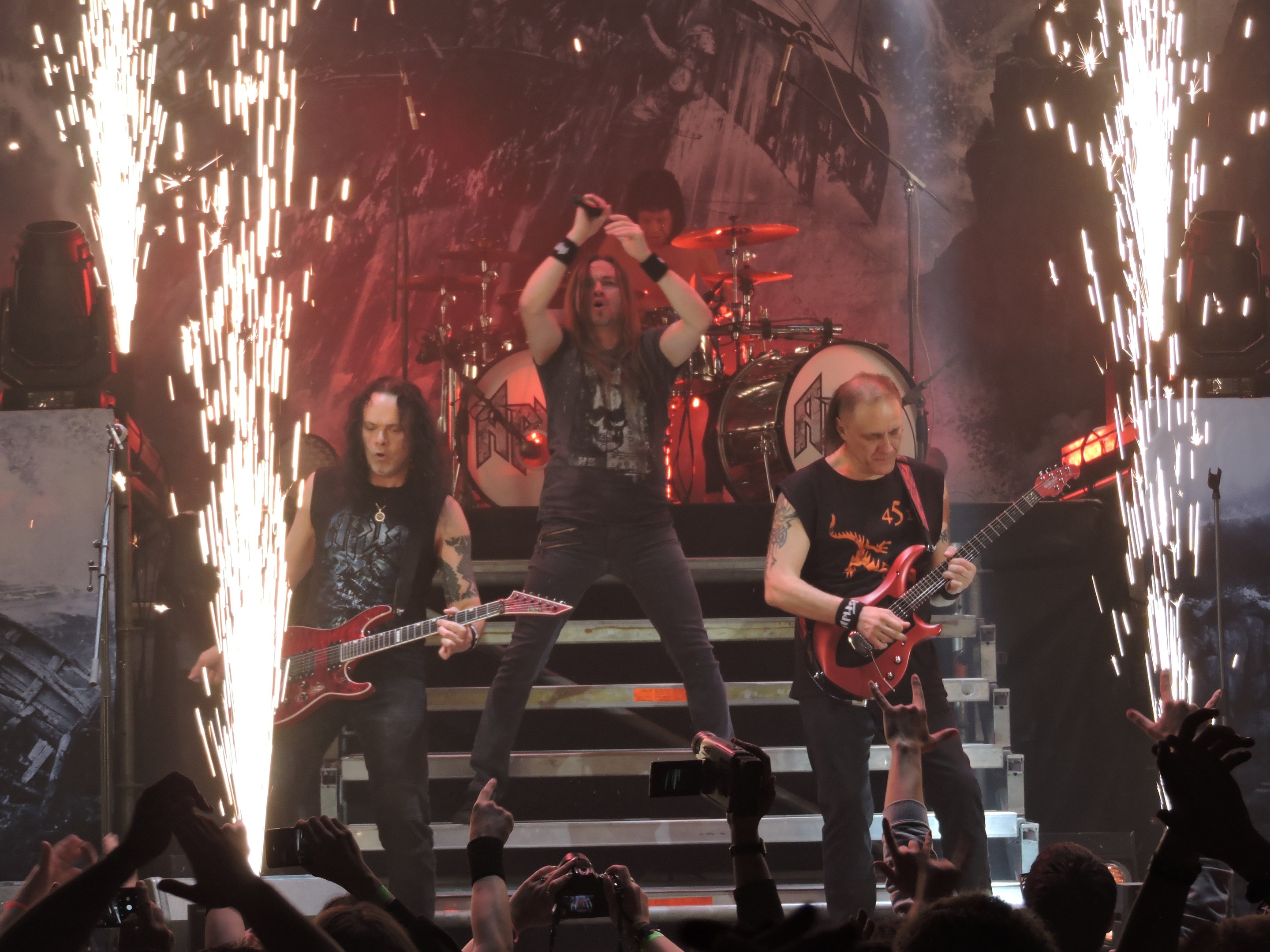 Eine Gruppe von Menschen, die auf einer Bühne bei einem Konzert mit Gitarren, einem Mikrofon und Trommeln spielen, während das Publikum das Ereignis mit Handys aufzeichnet.