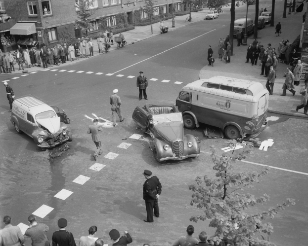 Schwarz-weiß-Bild eines Autounfalls an einer Straßenecke mit mehreren Fahrzeugen, darunter ein Lieferwagen, und einer Gruppe von Menschen in der Nähe, mit Gebäuden, Bäumen, Masten und Texttafeln im Hintergrund.