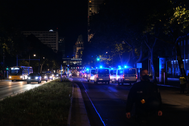 Eine nächtliche Straßen Szenerie in einer Stadt mit Polizeiwagen, Bussen, Passanten auf dem Gehweg, Gras, Bäumen, Gebäuden und beleuchteten Laternen.