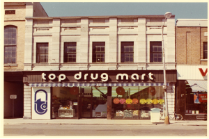 Top-Drogenmart-Geschäftsstelle an einer Stra√enecke mit einem Gebäude, einer Stra√enlampe, einem geparkten Fahrrad, einem Müllcontainer und einem bewölktem Himmel.