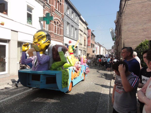 Eine Gruppe von Menschen auf einem Paradewagen, der mit bunten Spielzeugen geschmückt ist, umgeben von Gebäuden mit Fenstern, Texttafeln, Bäumen und einem klaren blauen Himmel.
