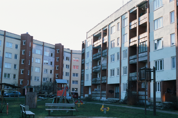 Ein Wohnhaus mit einem Spielplatz davor, mit Gras, Bänken, Schaukeln und anderen Gegenständen, mit Fahrzeugen, Bäumen und einem klaren blauen Himmel im Hintergrund.