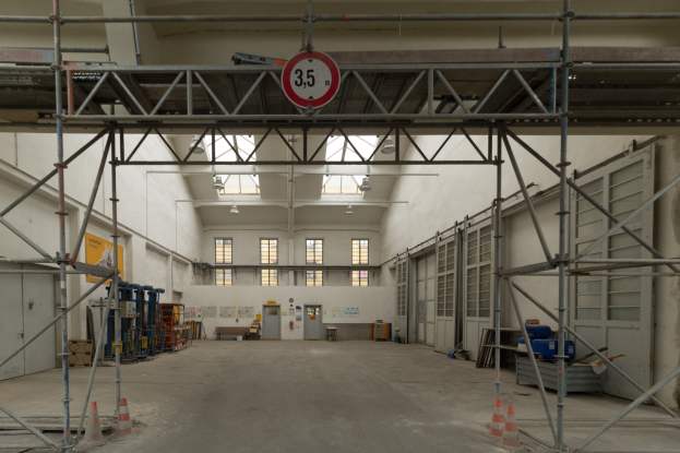 Ein großes Lager mit umfangreichen Gerüsten, Verkehrskegeln auf dem Boden, Metallstangen, ein Schild, verschiedene Maschinen, eine Tür, Fenster, eine Wand und ein Dach mit Deckenbeleuchtung.