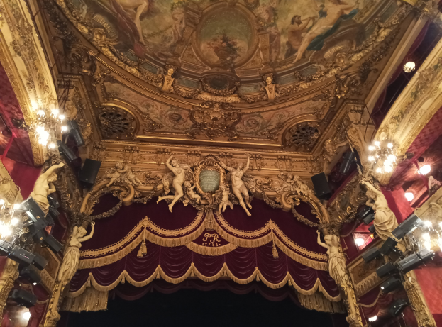 Theaterinnenraum mit roter Gardine, Kronleuchtern, Skulpturen, Lichtern und einer Deckenmalerei, mit einer Bühne und einer kleinen Gruppe von Menschen davor.