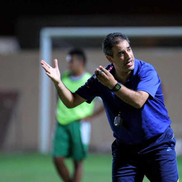 Ein Mann in einem blauen T-Shirt und blauen Hosen steht auf einem Fußballfeld und hält ein Mikrofon in der Hand, während er auf seine Spieler zeigt. Im Hintergrund sind eine weitere Person in einem grünen Kleid, ein Torpfosten und eine Wand zu sehen.