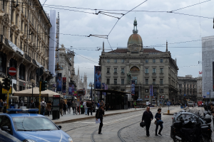 Eine belebte Stadtstraße mit einem geparkten Polizeiwagen, Fußgängern mit Taschen, fahrenden Fahrzeugen, hohen Gebäuden mit Fenstern, Bannern, Laternen, Ampeln und einem bewölkten Himmel.