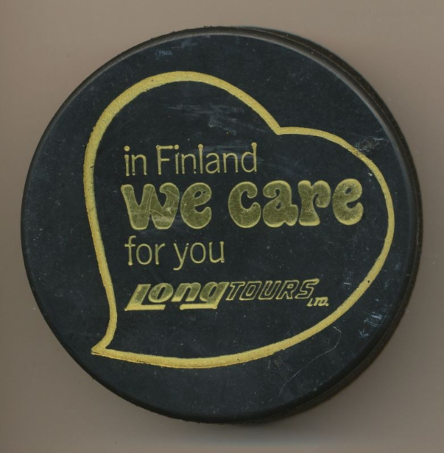 Schwarzer Hockey-Puck mit der Aufschrift "In Finland We Care For You Long Tours" auf cremefarbenem Hintergrund.