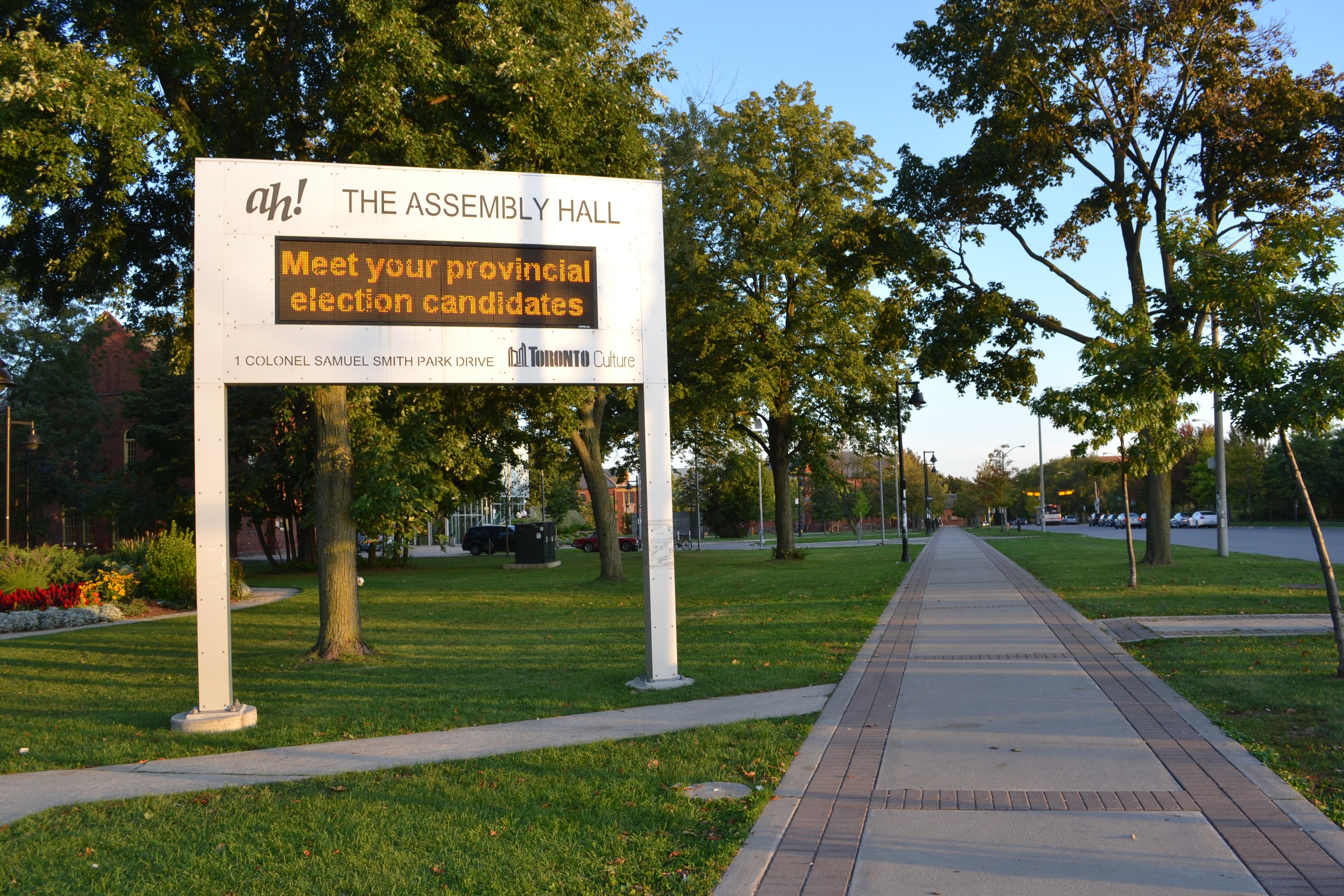 Rat auf Gras mit Text "Die Versammlungshalle - Treffen Sie Ihre Provincial Election Candidates" in der Nähe von Weg, Bäumen, Blumen, Straßenlaternen, Fahrzeugen, Gebäude und bewölktem Himmel.