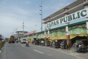 Eine belebte Stadtstraße mit Fahrzeugen, einem Geländer auf der linken Seite, Gebäuden, Polen, Lampen, Schildern und einigen Menschen unter einem bewölkten Himmel, mit einem Gebäude mit der Aufschrift 'Capas Public'.