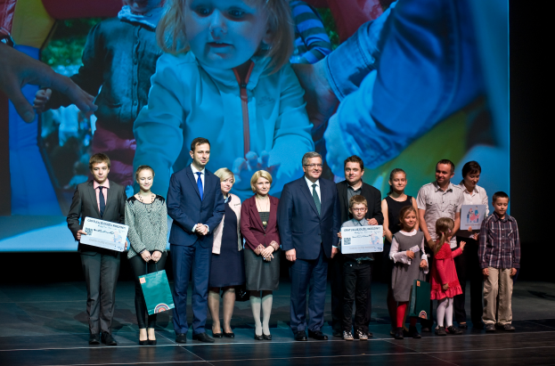 Eine Gruppe von Menschen auf einer Bühne, einige halten Taschen und Zertifikate, mit einem Bildschirm im Hintergrund, der ein Bild eines Kindes und anderer Menschen anzeigt.