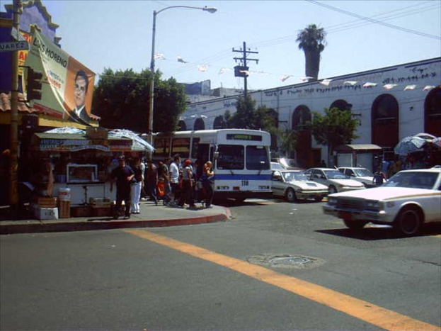 Eine belebte Straßenszene mit zahlreichen Fahrzeugen, Gebäuden, einem Stand, Bäumen und einem klaren Himmel.