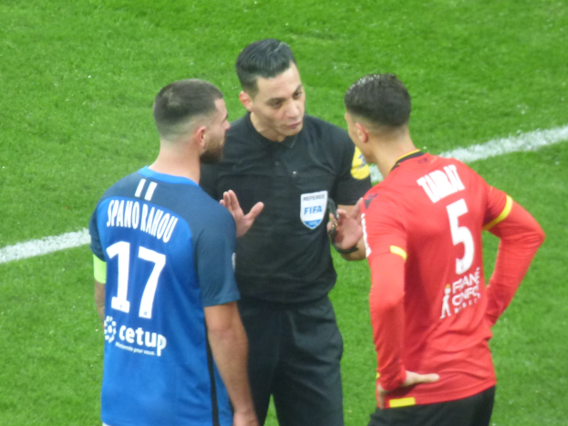 Schiedsrichter in schwarzem Hemd und Hose, der mit zwei Männern auf einem Fußballfeld spricht; ein Mann trägt ein blaues und rotes T-Shirt mit Text, beide tragen eine Brille.