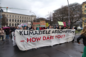 Eine Gruppe von Menschen marschiert mit einem Banner, auf dem "Klimaziele Abschaffen - Wie wagst du es?" steht, die Straße entlang, einige mit Schirmen und einer mit einem Megafon, mit Laternenmasten, Schildern, Bäumen, Gebäuden, einem Kran und einem klaren blauen Himmel im Hintergrund.