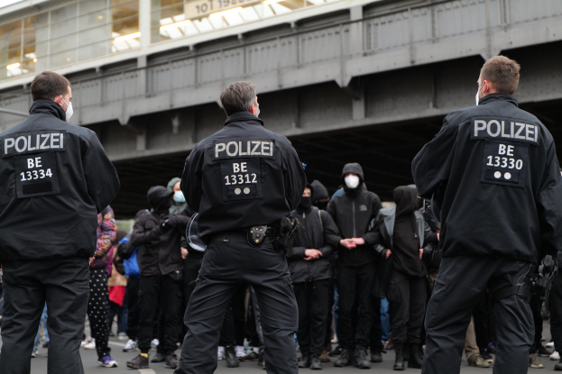 Eine Gruppe von Polizisten in Uniform steht vor einer Menge von Menschen in schwarzen Uniformen und Masken, mit einer Brücke und einem Gebäude im Hintergrund, was auf eine städtische Szenerie während einer Demonstration hindeutet.