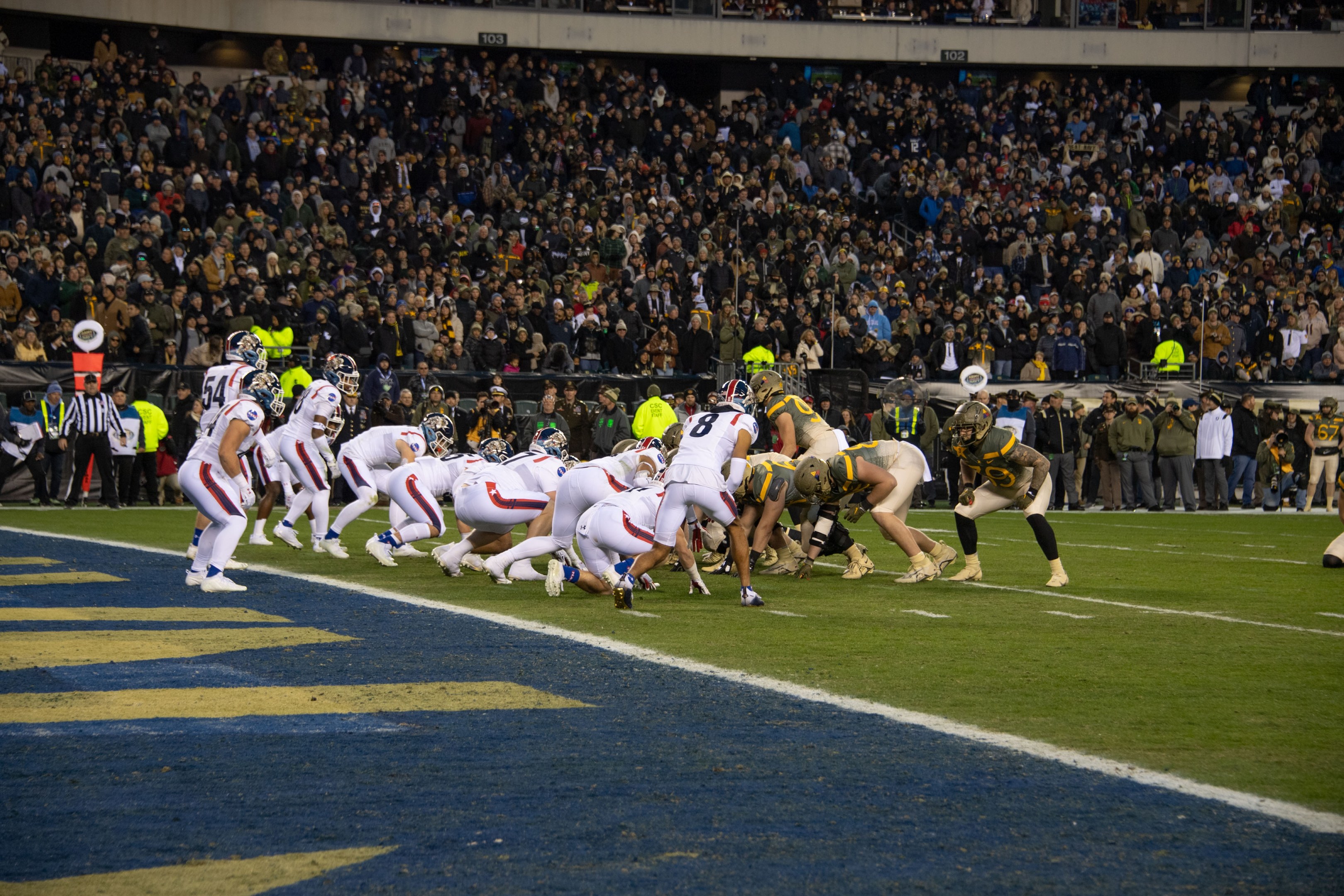 Ein Fußballspiel im Gange mit Spielern auf dem Boden in Sportbekleidung und Helmen, aus der Perspektive eines Stadionplatzes.