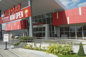 Ein Einkaufszentrum mit einem Schild, auf dem "City Mall Now Open" steht, umgeben von Pflanzen, Gras, einem Metallzaun, einer Gruppe von Menschen, einem Auto auf der Straße, Pfosten, einer Fahne, Lampen und einem bewölkten Himmel.