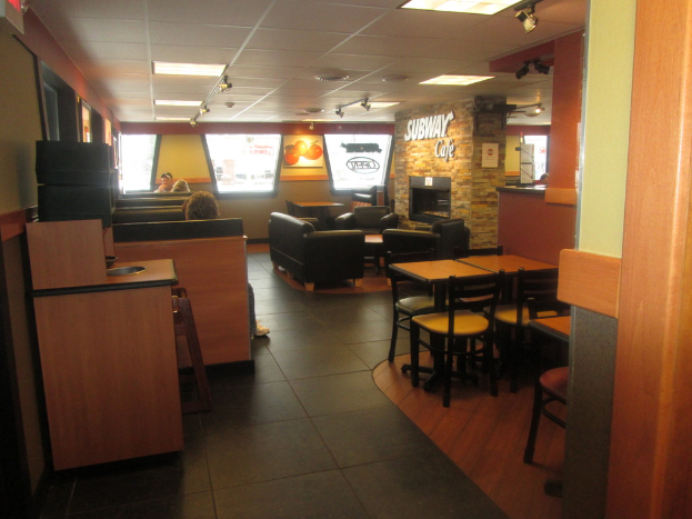 Ein U-Bahn-Café-Interieur mit Tischen, Stühlen, Sofas und vier Personen, eine Person trägt eine Mütze in der Nähe von Fenstern und Plakaten, ein Tisch in der rechten Ecke, Lampen, Überwachungskameras und eine Wand mit 'Subway Cafe' darauf.