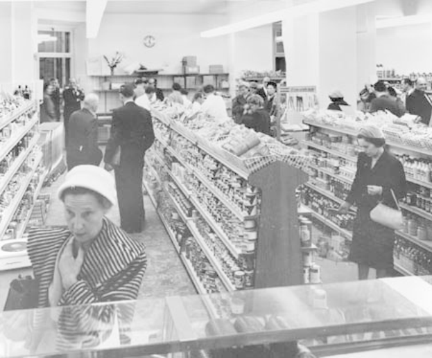Schwarzes und weißes Foto von Einkäufern in einem Supermarkt mit Lebensmittelregalen, Menschen mit Taschen, einer Wanduhr und Deckenleuchten.